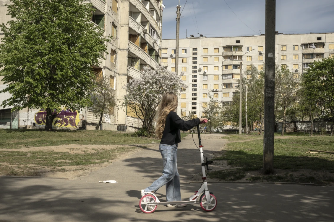 A girl rides a scooter against the backdrop of buildings damaged by shelling in Kharkiv. 